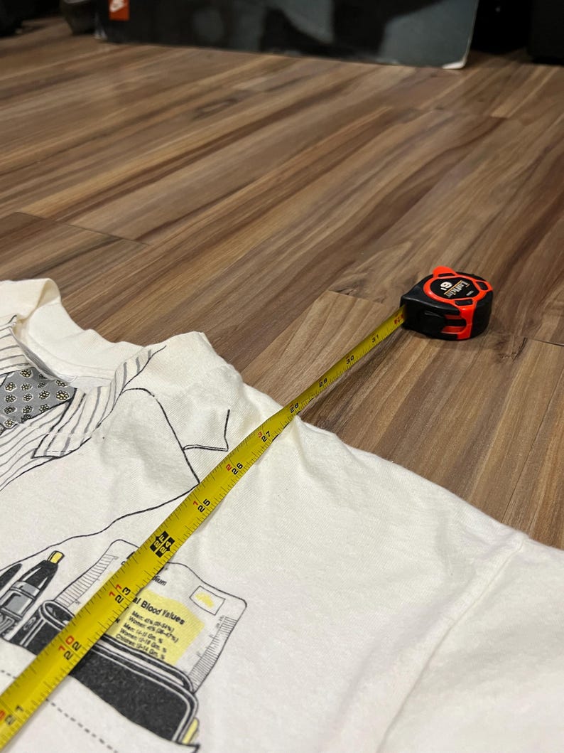 May include: A white t-shirt with a graphic print of medical equipment and text. A yellow measuring tape extends across the shirt, showing measurements in inches. The tape measure is black and red.