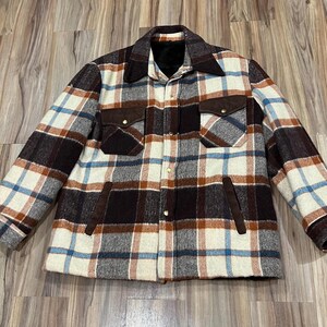 May include: A long-sleeve button-up jacket featuring a plaid pattern in cream, dark brown, light blue, and rust orange. The garment includes a dark brown collar, two chest pockets with flaps, two side pockets, and gold-toned metal snap buttons.