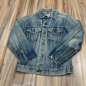 May include: A blue denim jacket with a distressed finish, featuring a pointed collar, long sleeves, two button-flap chest pockets, a front button closure, and frayed edges along the cuffs, hem, and collar, with a small red tab on the left pocket.