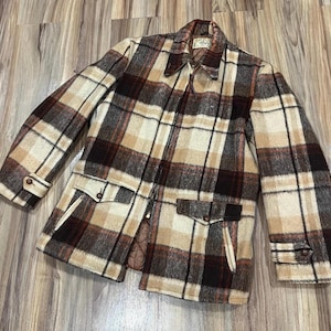 Könnte beinhalten: Eine braune, beige und schwarze karierte Jacke mit Reißverschluss. Die Jacke hat zwei große Taschen auf der Vorderseite und ein gestepptes Futter.