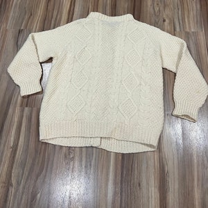 May include: A cream-colored knitted sweater with a cable knit pattern. The sweater has a high neck and long sleeves.