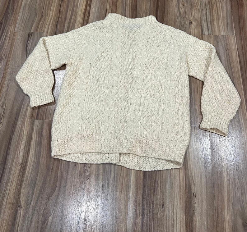 May include: A cream-colored knitted sweater with a cable knit pattern. The sweater has a high neck and long sleeves.