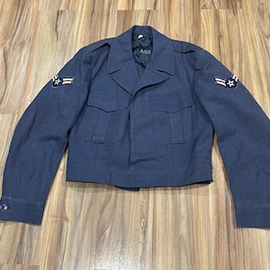 May include: A blue Air Force jacket with two front pockets and patches on the sleeves. The jacket has a button closure and a collar. The jacket is made of a durable fabric and is likely part of a uniform.