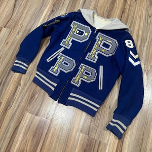May include: A blue varsity jacket with a gray hood and white and blue stripes on the sleeves and bottom. The jacket has a zipper closure and features large gray felt letters that spell "PP" with the word "Valley" written in yellow on each letter. The jacket also has a yellow number "8" and a chevron on the right sleeve.