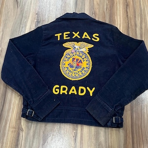 Puede incluir: Una chaqueta de pana azul oscuro con texto bordado en amarillo que dice TEXAS y GRADY. Centrado en la espalda hay un emblema circular de FFA Vocational Agriculture con un diseño de águila, búho y arado en hilo amarillo, azul y rojo.