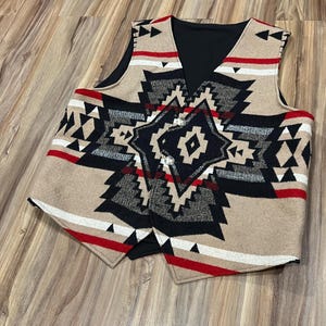 Puede incluir: Un chaleco beige, negro, rojo y blanco con un diseño geométrico. El chaleco tiene cuello en V y dos botones plateados. El patrón incluye triángulos, diamantes y rayas. El chaleco está hecho de un material grueso y tejido.