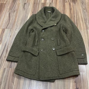 May include: An olive green double-breasted coat featuring a wide shawl collar, long sleeves with buttoned cuffs, and two large front flap pockets. The coat is constructed from a textured wool-like fabric and includes six dark buttons on the front.