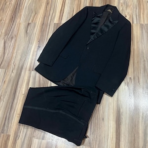 May include: A black suit set, including a blazer and trousers, is displayed on a wooden surface. The blazer has a satin lapel and a single-button closure. The trousers are neatly folded, showcasing a classic, formal style.