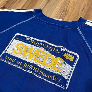 May include: A blue sweatshirt with white stripes on the sleeves and a yellow and white graphic print of a Minnesota license plate that says "SWEDE land of 10,000 Swedes".