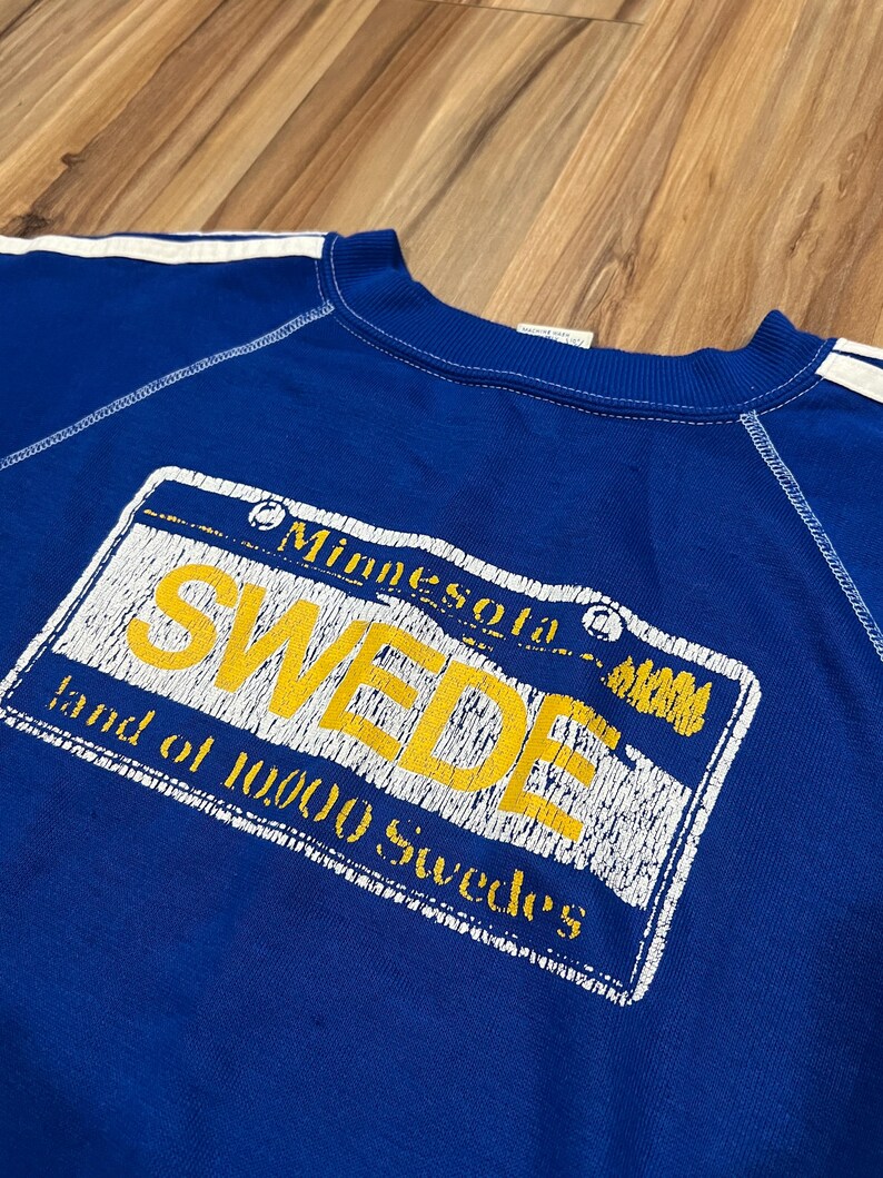 May include: A blue sweatshirt with white stripes on the sleeves and a yellow and white graphic print of a Minnesota license plate that says "SWEDE land of 10,000 Swedes".