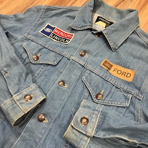 May include: A light blue denim jacket featuring a collar, long sleeves with buttoned cuffs, and a front button closure. The jacket includes a rectangular patch with the text Ford, Mercury, and Lincoln, and a tan patch with the text Ford.