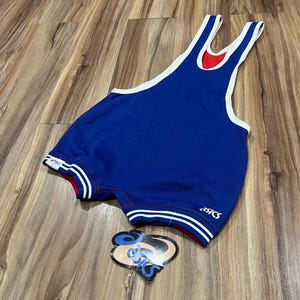 May include: Blue wrestling singlet with white and red trim. The singlet has a red interior and white accents around the neck and arm openings. The Asics logo is visible on the leg. A tag is attached.