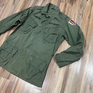 Peut inclure: Une veste de terrain de style militaire vert olive dotée de quatre grandes poches avant à rabats boutonnés, d'un col pointu et de manches longues à poignets boutonnés. La poitrine porte l'inscription U.S. ARMY et un écusson coloré est sur l'épaule.