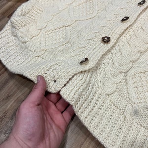 May include: A cream-colored knitted cardigan with a cable knit pattern and button closure. The cardigan is made of a soft, warm material and has a relaxed fit.