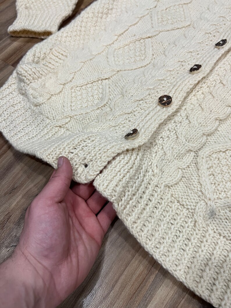 May include: A cream-colored knitted cardigan with a cable knit pattern and button closure. The cardigan is made of a soft, warm material and has a relaxed fit.