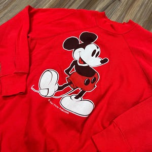 May include: A red sweatshirt featuring a large graphic of Mickey Mouse in his classic pose. Mickey is depicted in black, white, and red, with the words "Walt Disney Productions" printed below. The sweatshirt has long sleeves.