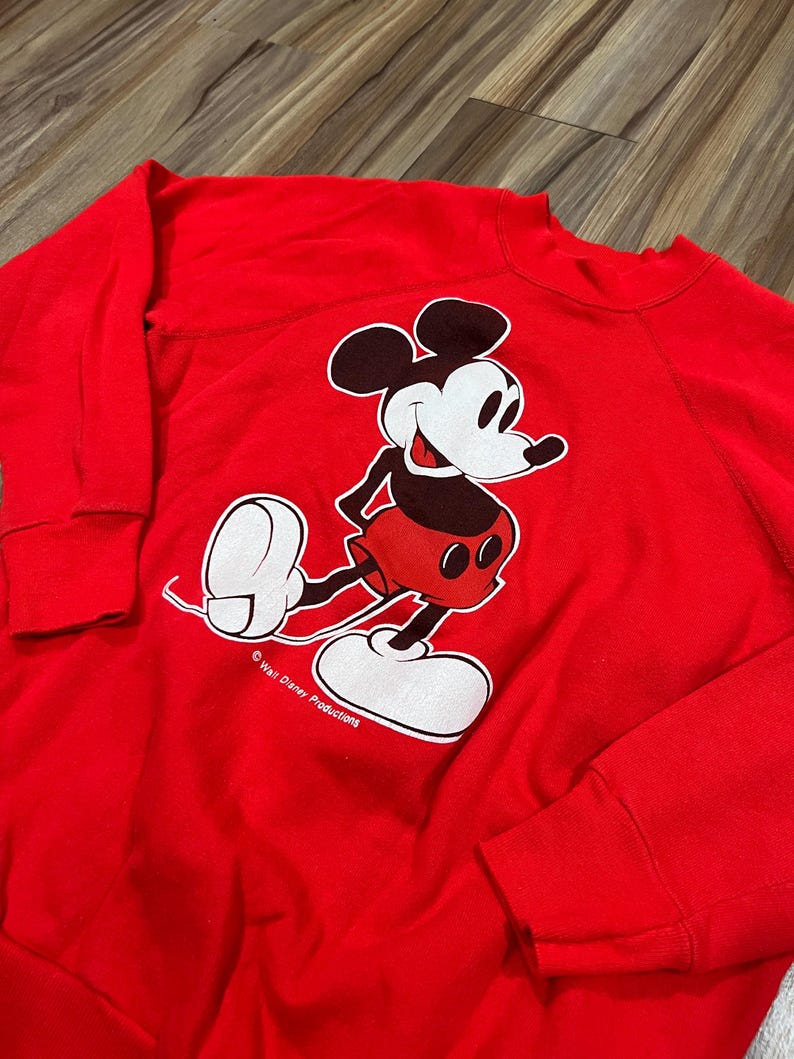 May include: A red sweatshirt featuring a large graphic of Mickey Mouse in his classic pose. Mickey is depicted in black, white, and red, with the words "Walt Disney Productions" printed below. The sweatshirt has long sleeves.