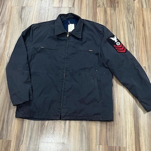 May include: A dark navy blue utility jacket featuring a full-length front zipper, a pointed collar, and two horizontal zippered chest pockets. The left sleeve displays a black patch with a white eagle emblem and three red chevron stripes.