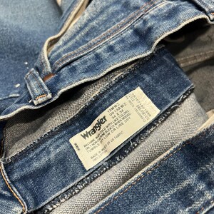 Pu&ograve; includere: Jeans in denim blu con un'etichetta interna bianca riportante il logo Wrangler e testo che include 100% cotone, prodotto in Messico con tessuto statunitense, e taglia 34 x 34. I jeans presentano rivetti color rame e cuciture visibili in vita.