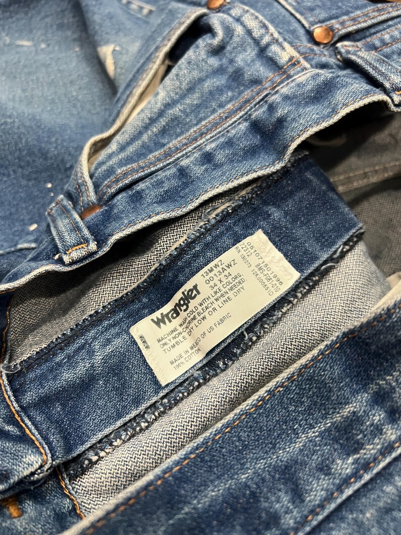 Pu&ograve; includere: Jeans in denim blu con un'etichetta interna bianca riportante il logo Wrangler e testo che include 100% cotone, prodotto in Messico con tessuto statunitense, e taglia 34 x 34. I jeans presentano rivetti color rame e cuciture visibili in vita.