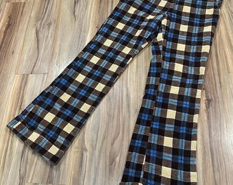 May include: A pair of plaid pants in a blue, brown, and cream color scheme. The pants have a wide leg and a black waistband. The fabric appears to be a soft, textured material. The pants are laid out on a wooden surface.