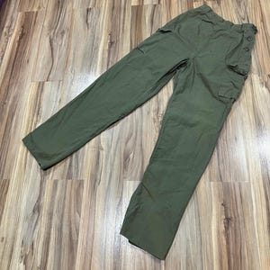 May include: Olive green cargo pants with multiple pockets and button details. The pants are made of a durable fabric and have a relaxed fit. The pants are laid out on a wooden surface.