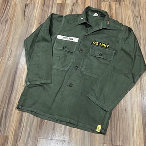 Peut inclure: Chemise militaire vert olive à manches longues, deux poches de poitrine et fermeture boutonnée. Comporte une étiquette nominative "SHULICK" et un écusson "U.S. ARMY". La chemise a un col et des épaulettes.