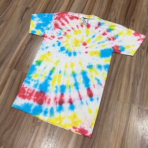 May include: A white t-shirt with a tie-dye pattern in shades of red, blue, and yellow. The shirt has a circular pattern with a white center.