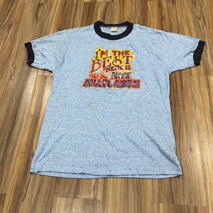 Puede incluir: Camiseta de color azul claro con ribetes negros en el cuello y las mangas. La camiseta presenta el texto "I'M THE BEST THESE IS BUT I'M NOT AVAILABLE" en un diseño colorido de estilo vintage. La camiseta está tendida sobre una superficie de madera.