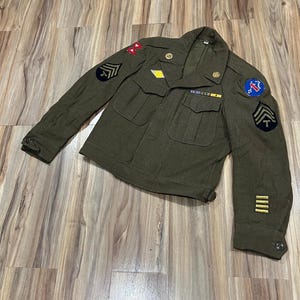 May include: An olive drab wool military jacket featuring two chest pockets, notched lapels, and gold-tone buttons. The sleeves and shoulders display various embroidered patches, including rank insignia, unit emblems, and four horizontal service stripes.