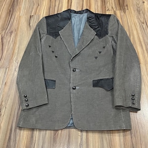 May include: A corduroy blazer in a muted grey-green colour with black leather accents on the shoulders and pockets. The blazer has a three-button closure and decorative stitching. The lining is a light blue colour.