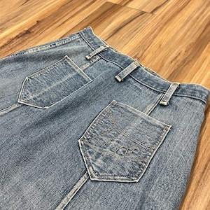 May include: Close-up of light blue denim jeans. The jeans feature a back pocket with a stitched design and a seam down the center. The jeans are on a wooden surface.