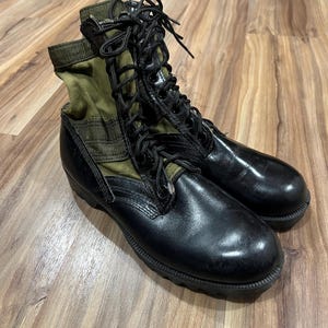 May include: Pair of black combat boots with olive green canvas uppers and black laces. The boots have a glossy finish and are set on a wooden floor.