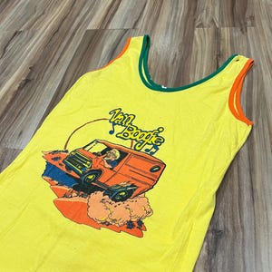 以下が含まれることがあります： 黄色のタンクトップ、緑とオレンジのトリム付き。前面には、運転手、煙、そして「Van Booge」のテキストが描かれたオレンジ色のバンのグラフィックが描かれています。タンクトップは柔らかく、軽量な素材で作られています。