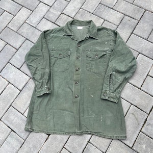 Op de afbeelding: Een groen, lang mouwig overhemd met een knoopsluiting en twee grote zakken. Het overhemd heeft een vervaagde uitstraling en lijkt vintage te zijn.