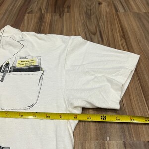 May include: White t-shirt with a pocket design featuring a pen and a wallet. The pocket also contains a card with the text "Ketzel Normal Blood Values". A yellow measuring tape is placed across the shirt, showing measurements from 17 to 27 inches.