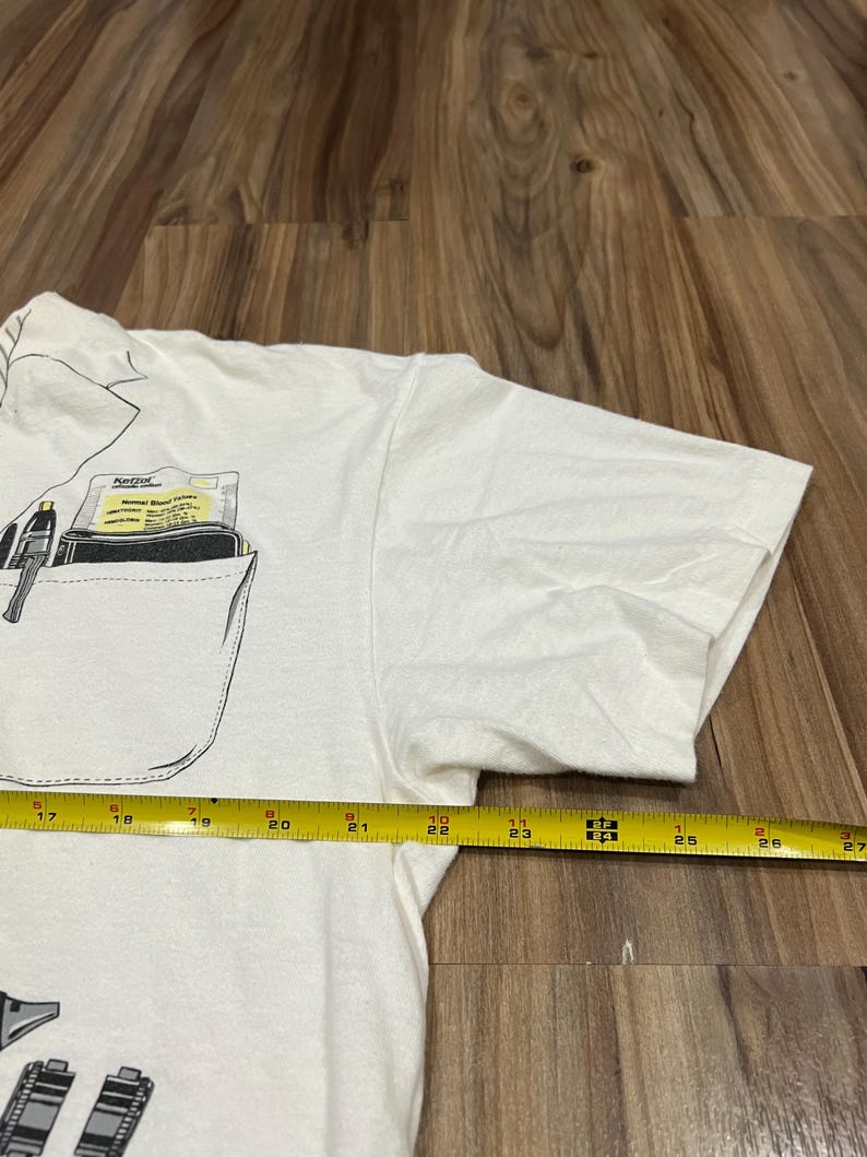 May include: White t-shirt with a pocket design featuring a pen and a wallet. The pocket also contains a card with the text "Ketzel Normal Blood Values". A yellow measuring tape is placed across the shirt, showing measurements from 17 to 27 inches.