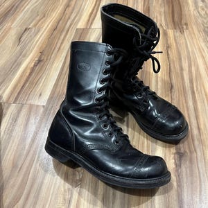 May include: Pair of black leather combat boots with a glossy finish. The boots have a lace-up design with metal eyelets and a rounded toe. The boots are on a wooden surface.