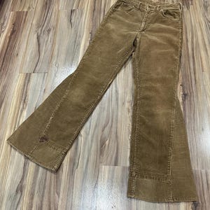 May include: Brown corduroy pants with a flared leg design. The pants have a classic five-pocket style and a button and zipper closure. The corduroy fabric has a ribbed texture and a vintage look.