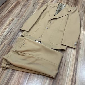 Puede incluir: Un traje color beige, compuesto por una americana y un pantalón a juego, se muestra sobre una superficie de madera. La americana tiene dos bolsillos delanteros y cierre de botones. El pantalón está doblado junto a la chaqueta.