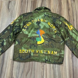 May include: Camouflage-patterned jacket with the text "LONG THANH NORTH SOUTH VIET NAM 1968 1969". The jacket features a map graphic with colorful accents and gold embroidery on the sleeves.