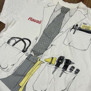 May include: White t-shirt with a doctor's coat design. The shirt features a printed tie, pockets with medical tools, and the word "Hawaii" in red. The design includes a stethoscope and a belt.