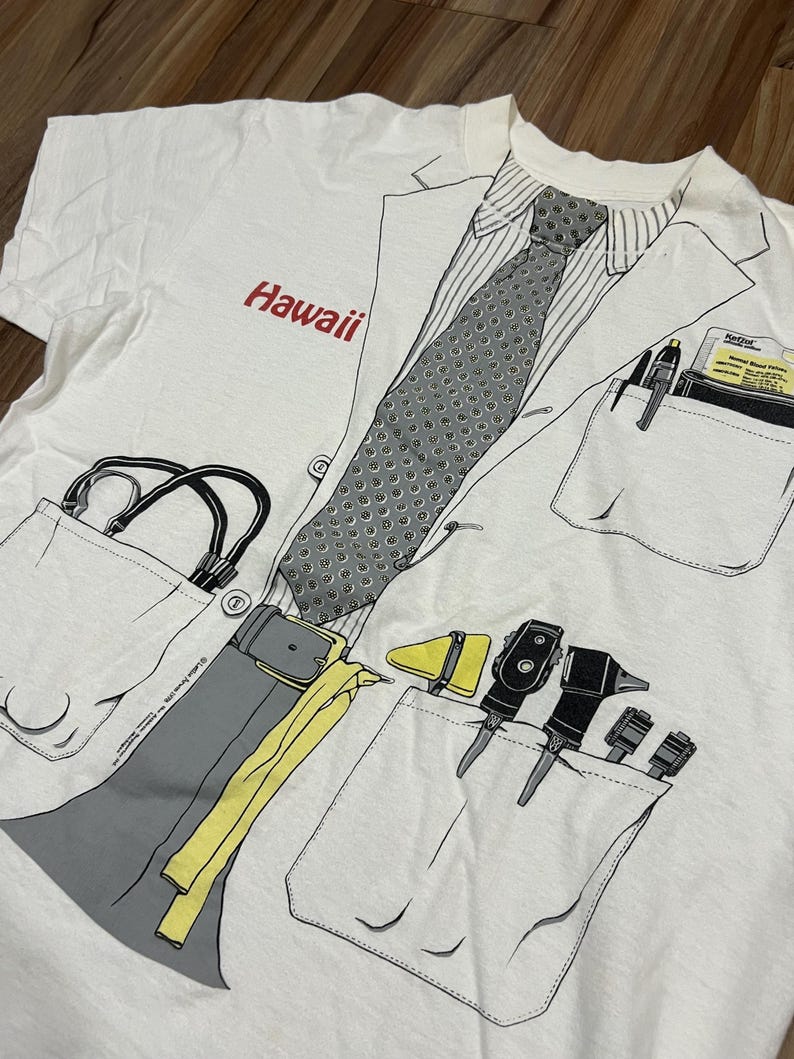 May include: White t-shirt with a doctor's coat design. The shirt features a printed tie, pockets with medical tools, and the word "Hawaii" in red. The design includes a stethoscope and a belt.