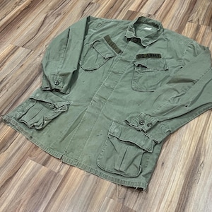 May include: An olive green military jacket with multiple pockets and a button-down front. The jacket has a collar and the words "U.S. ARMY" are visible on a patch. The jacket is made of a durable fabric and has a worn appearance.