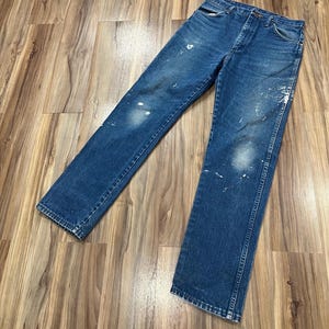 Pu&ograve; includere: Un paio di jeans in denim blu con classico design a cinque tasche, chiusura con bottoni e taglio a gamba dritta. Il tessuto presenta un lavaggio medio con visibili schizzi di vernice bianca e zone sbiadite su ginocchia e cosce.