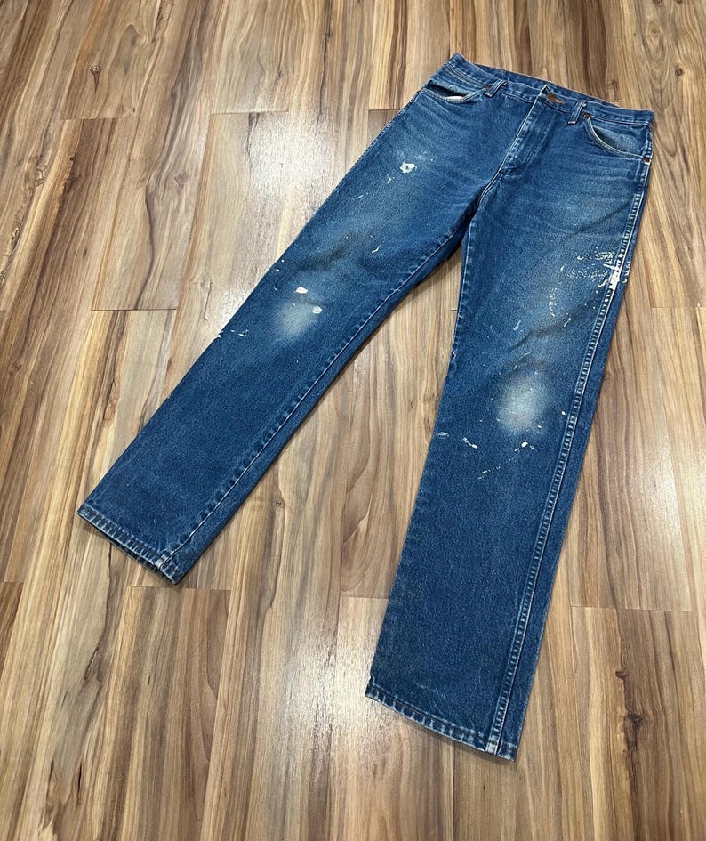 Pu&ograve; includere: Un paio di jeans in denim blu con classico design a cinque tasche, chiusura con bottoni e taglio a gamba dritta. Il tessuto presenta un lavaggio medio con visibili schizzi di vernice bianca e zone sbiadite su ginocchia e cosce.