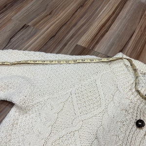 May include: A white knitted cardigan with a cable knit pattern and wooden buttons. The cardigan is laid flat on a wooden surface and measured with a tape measure.