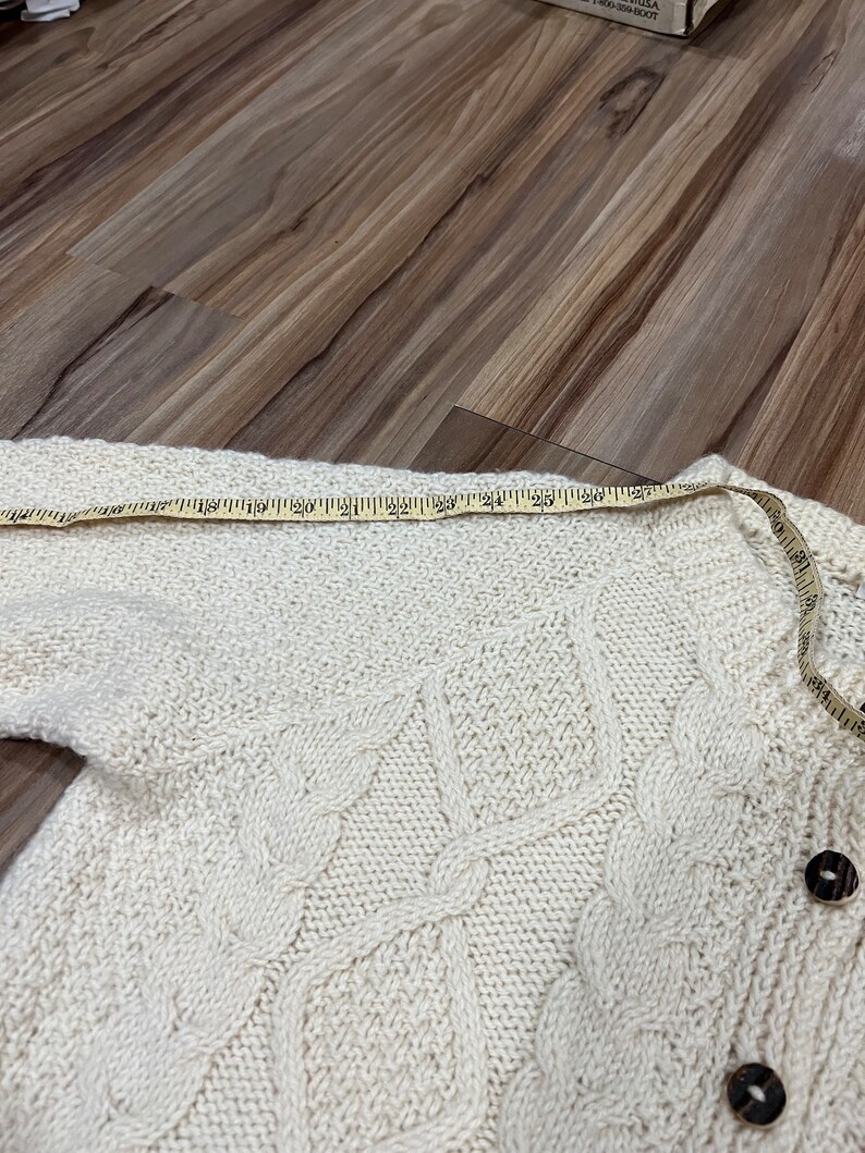 May include: A white knitted cardigan with a cable knit pattern and wooden buttons. The cardigan is laid flat on a wooden surface and measured with a tape measure.