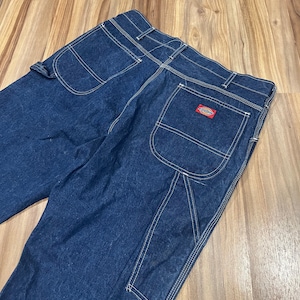 May include: Dark blue denim work pants with multiple pockets and white stitching. The back pocket features a red and white Dickies logo patch. The pants are made of a durable material.