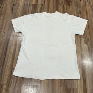 May include: A plain white t-shirt with short sleeves, laid flat on a wooden surface. The shirt is made of a soft, likely cotton material. The t-shirt is a basic, versatile garment.